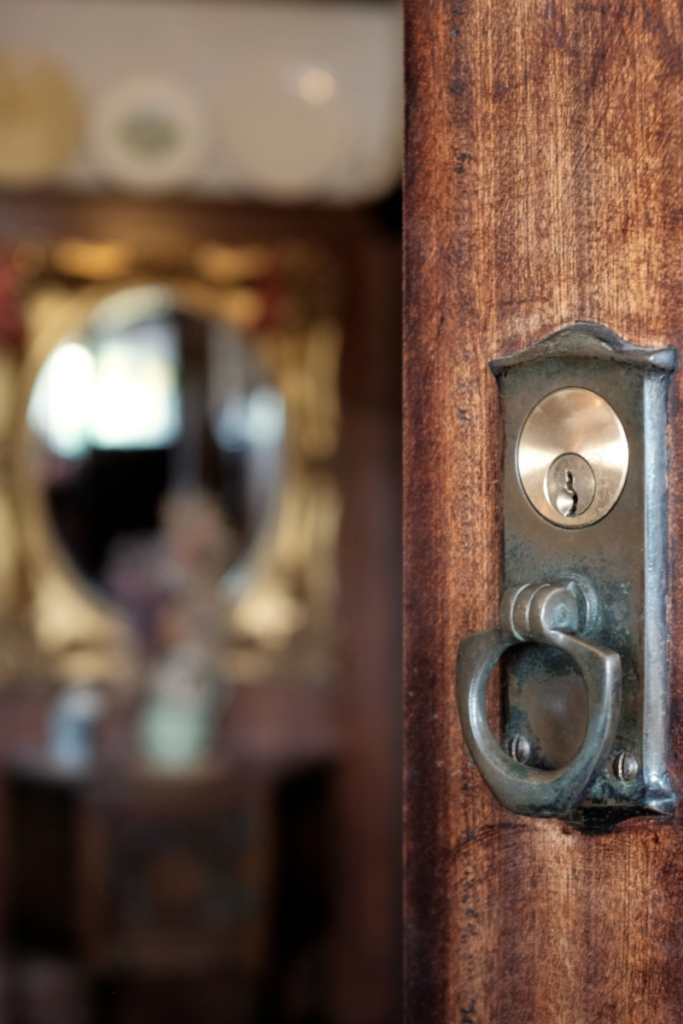 Picture of an old-fashioned rustic looking door lock with a knocker on the left side of the door in focus, while the background shows an out of focus circular wall mirror.
